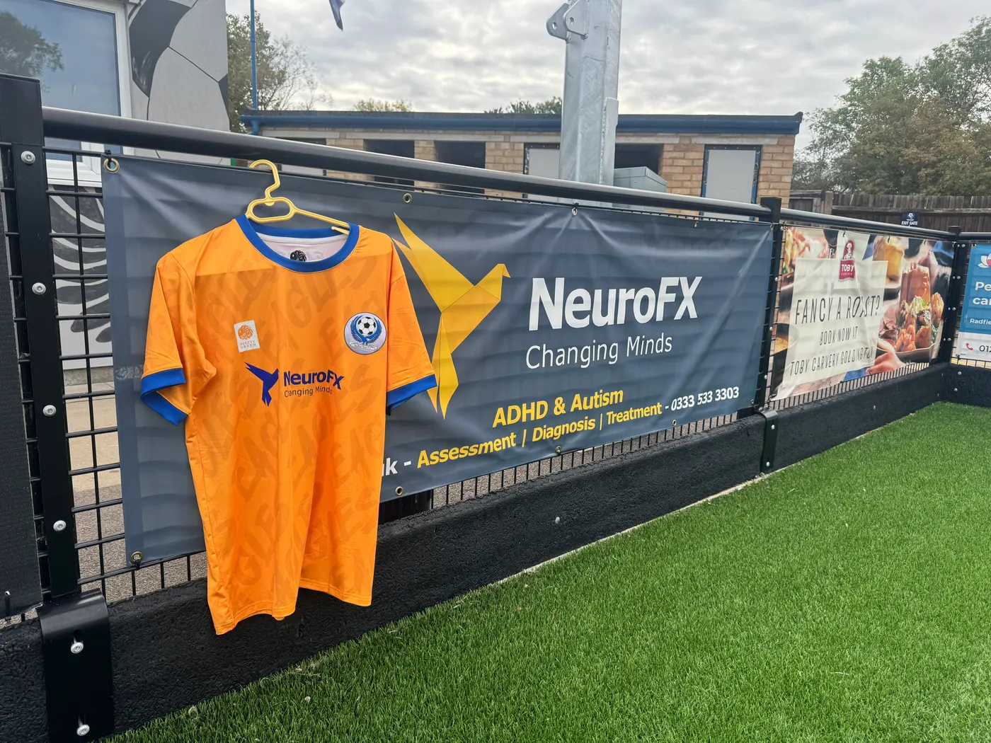 The amber and blue Bedford Town FC Academy shirt with the NeuroFX logo on the front, hanging beside the NeuroFX pitchside banner at the ground