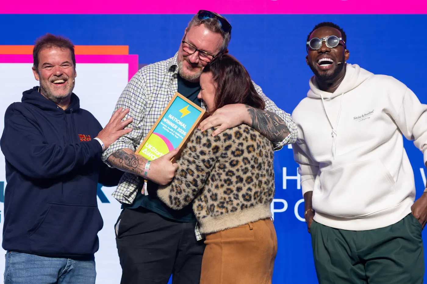 Tina and Paul hugging on stage while holding the National Winner 2026 certificate, the hosts laughing beside them