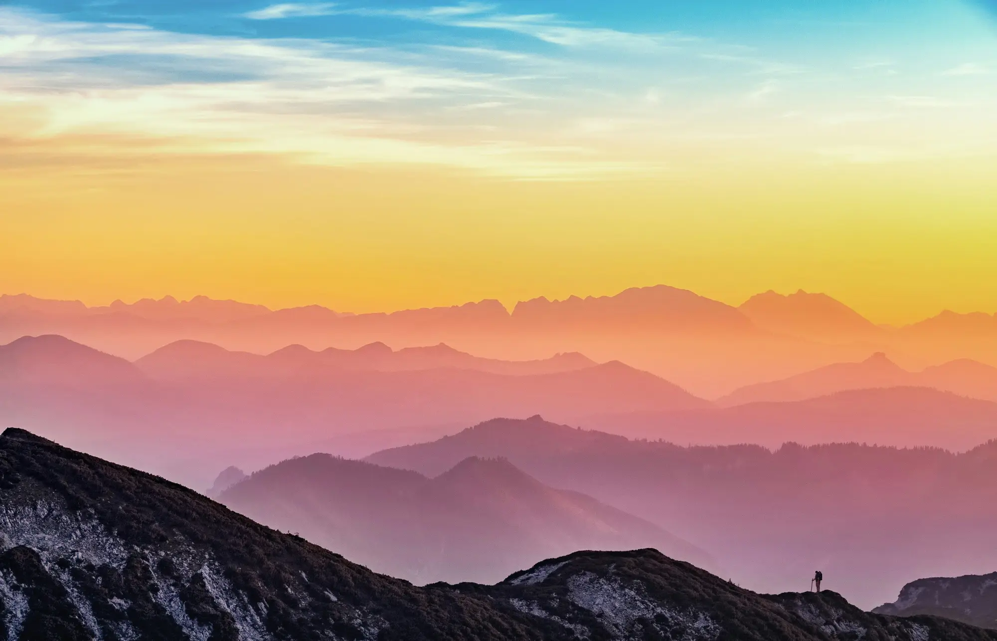 A lone walker on a ridge looking out over layers of distant hills in soft sunrise colours