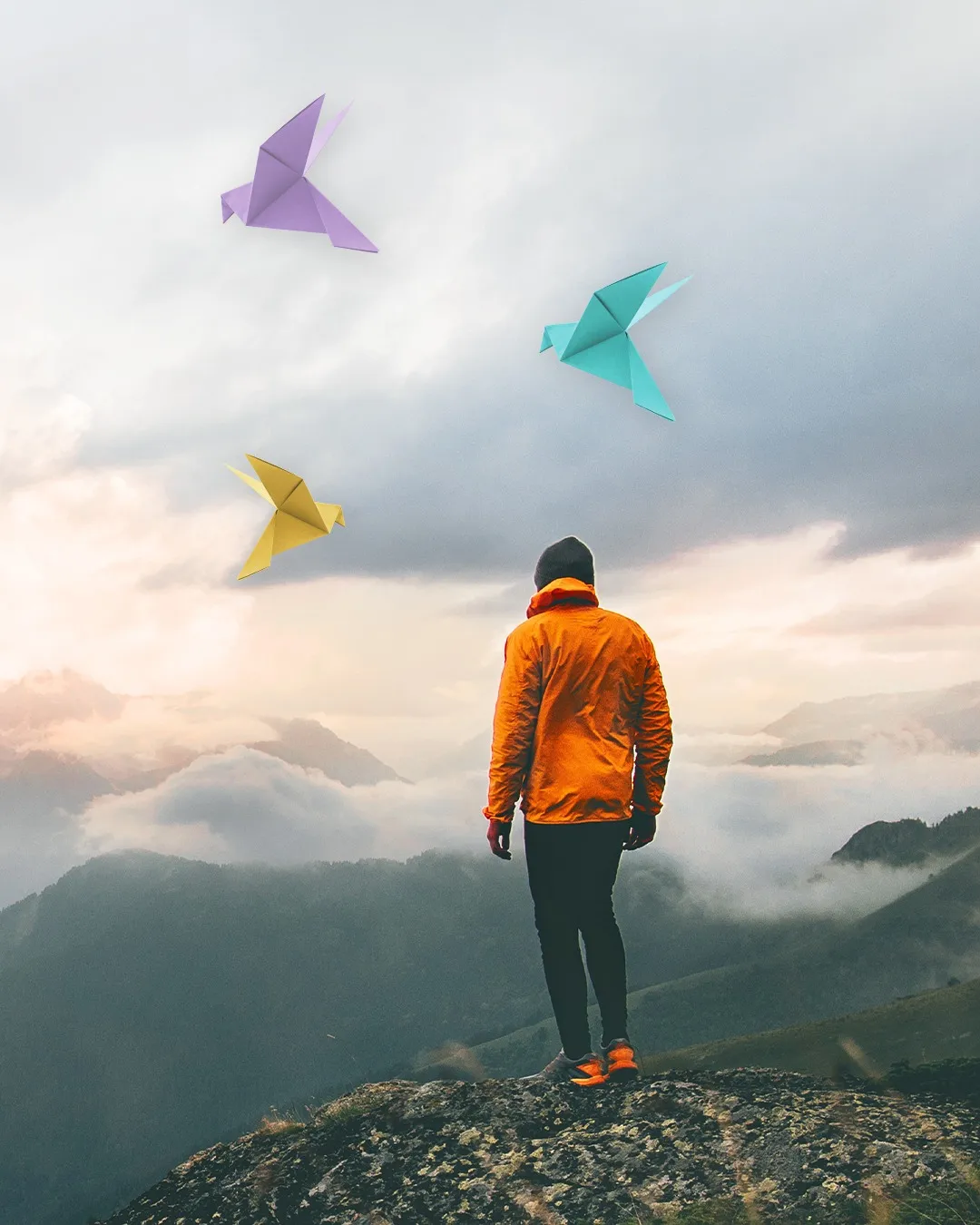 A person standing on a mountain ridge looking up at origami birds against a soft sky